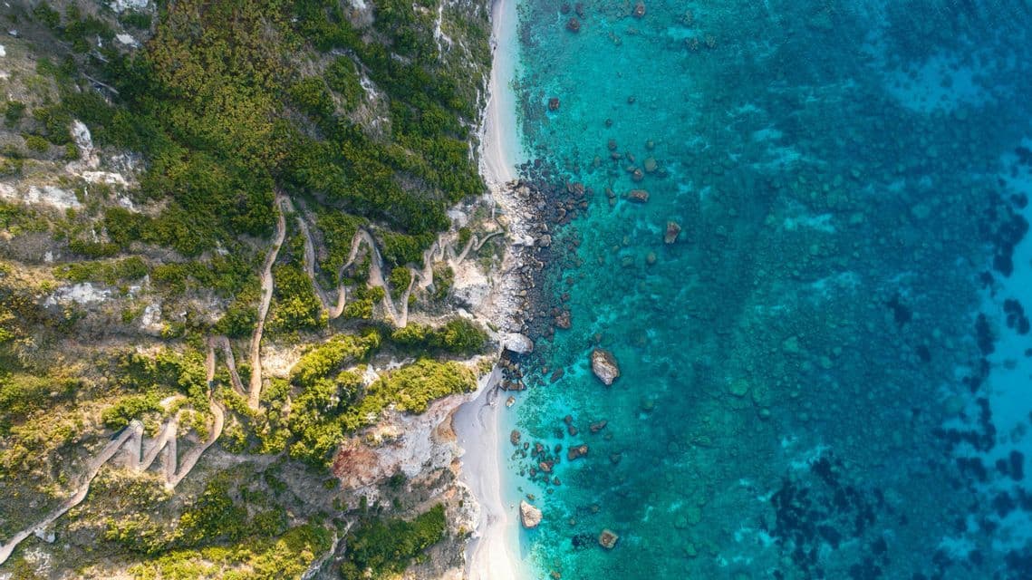 Una vista aerea dall'alto di un sentiero tortuoso che scende da una scogliera verde e lussureggiante fino a una spiaggia sabbiosa accanto a un mare turchese.