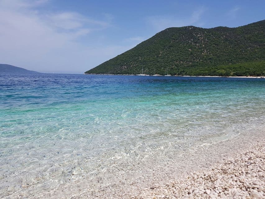 Una spiaggia di ciottoli bianchi con acqua cristallina e turchese e una grande collina boscosa sullo sfondo sotto un cielo blu.