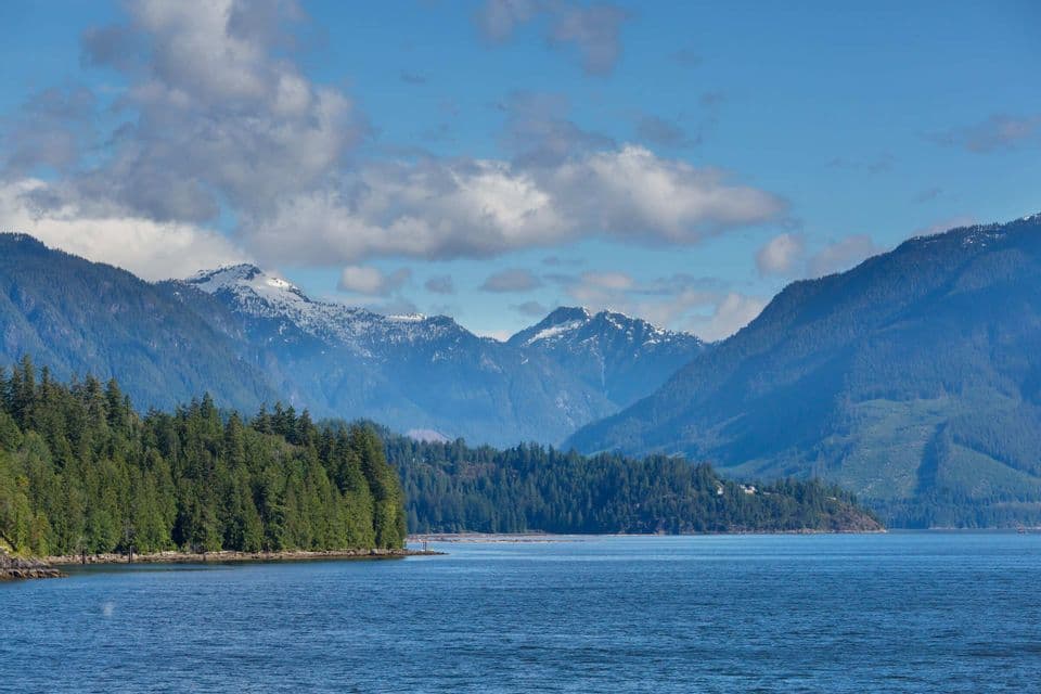 Ein Gewässer vor einem dichten Kiefernwald, mit einer Kette schneebedeckter Berge im Hintergrund unter einem teils bewölkten Himmel.