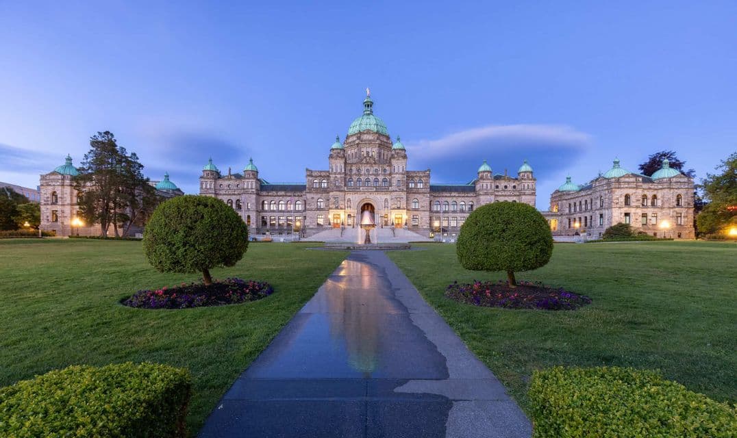 Ein nasser Steinweg führt in der Dämmerung zu einem großen, beleuchteten Gebäude mit Kuppeln, eingebettet in einen gepflegten Rasen mit runden Bäumen.