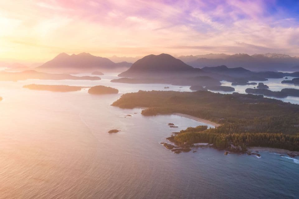 Luftaufnahme eines nebligen Archipels mit bewaldeten Inseln und fernen Bergen unter einem farbenprächtigen Sonnenaufgangshimmel.