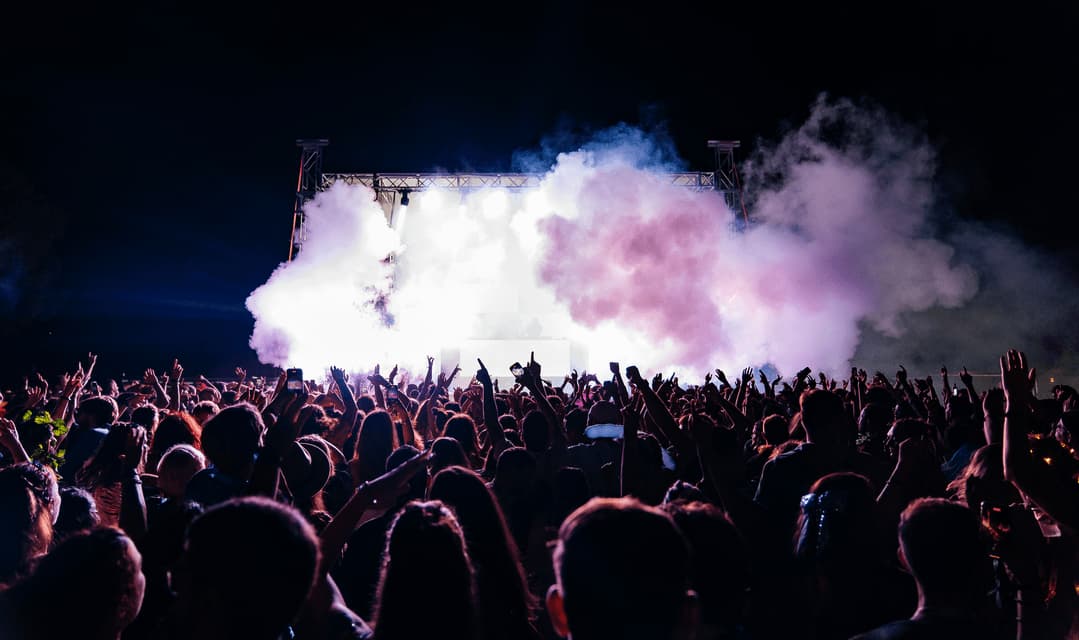 Una grande folla con le mani alzate è di fronte a un palco da concerto illuminato a giorno, con effetti di fumo viola e bianco, di notte.
