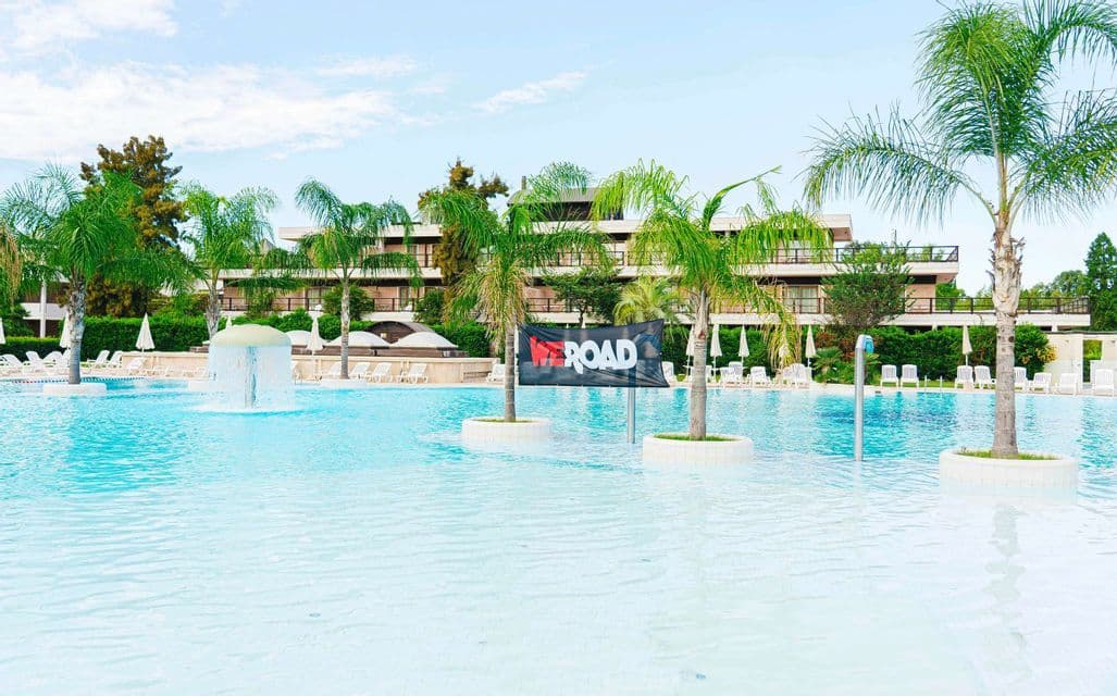 Un banner WeRoad è appeso tra due palme in una grande piscina di un resort, con un edificio alberghiero sullo sfondo.