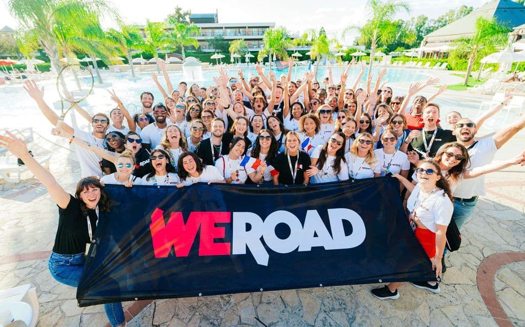 Un grande gruppo WeRoad esulta e saluta per una foto tenendo un grande striscione a bordo piscina di un resort.