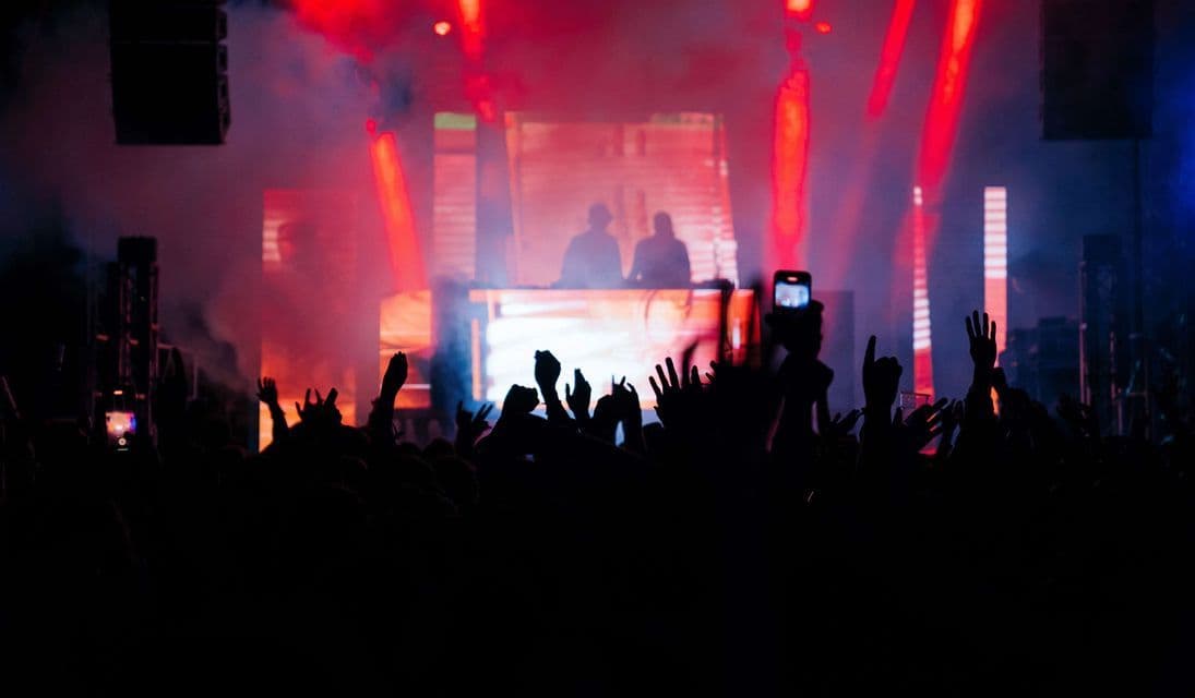 Sagome di una folla con le mani alzate a un concerto, davanti a due DJ su un palco con luci rosse e fumo.