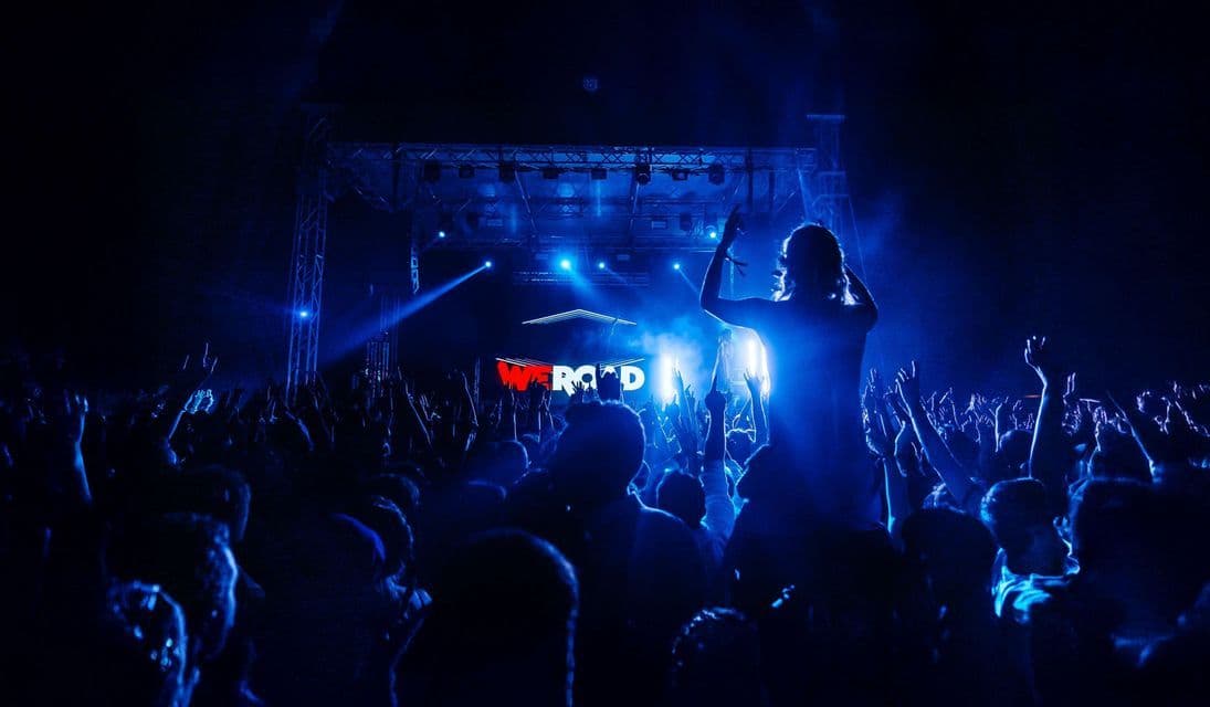 Una folla a un concerto WeRoad, sagomata dalle luci blu del palco con le mani alzate.