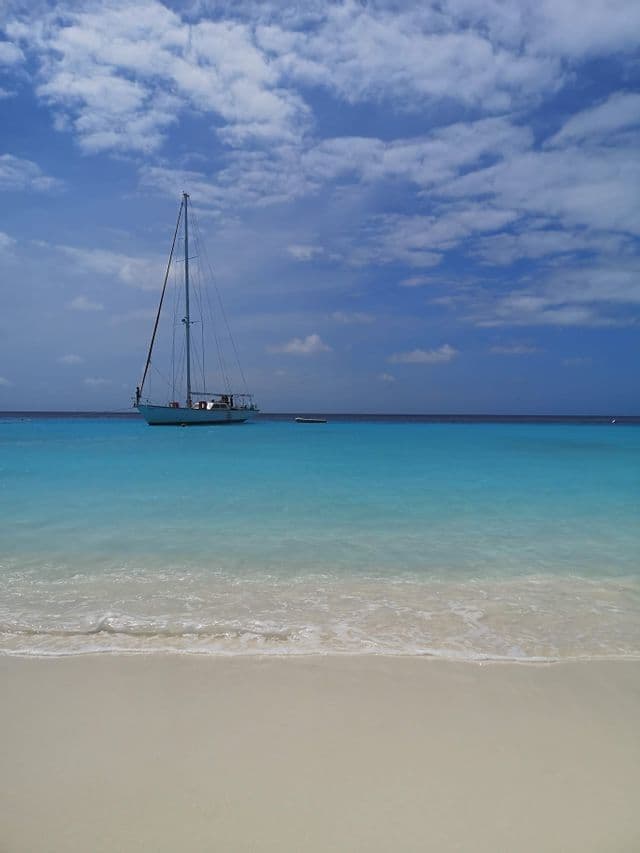 Una barca a vela ancorata in acque calme e turchesi, vista da una spiaggia di sabbia bianca sotto un cielo parzialmente nuvoloso.
