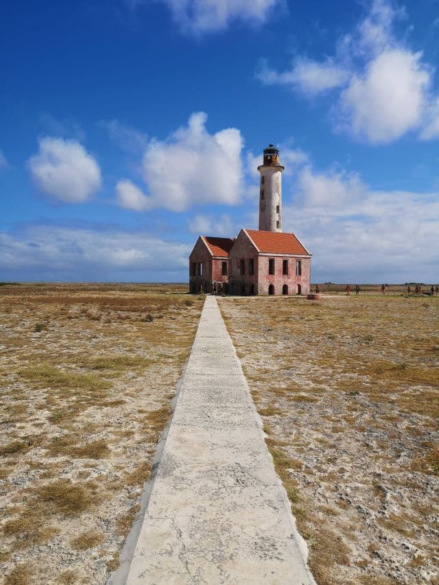 Un sentiero in cemento conduce a un vecchio faro in un campo arido sotto un cielo azzurro nuvoloso, con un viaggio di gruppo WeRoad nelle vicinanze.