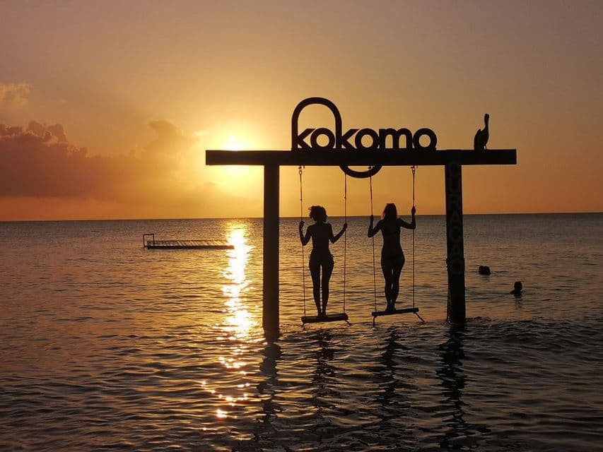 Due donne in silhouette si stagliano su altalene in mare, sullo sfondo di un tramonto dorato.