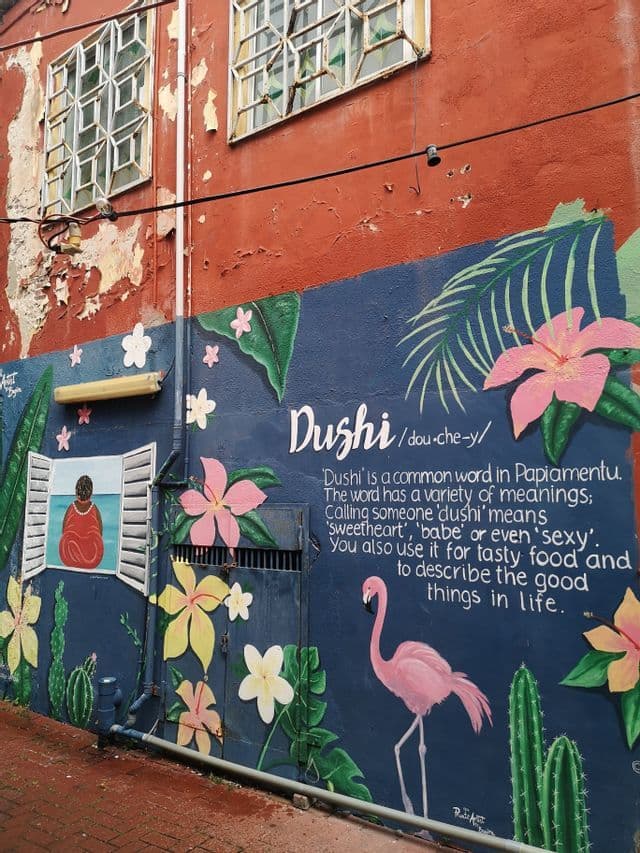 Un murale colorato su una parete di un edificio che raffigura un fenicottero rosa, fiori tropicali e la parola 'Dushi'.