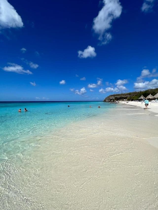 Una spiaggia di sabbia bianca con acque calme, limpide e turchesi dove le persone nuotano sotto un cielo azzurro intenso con nuvole.