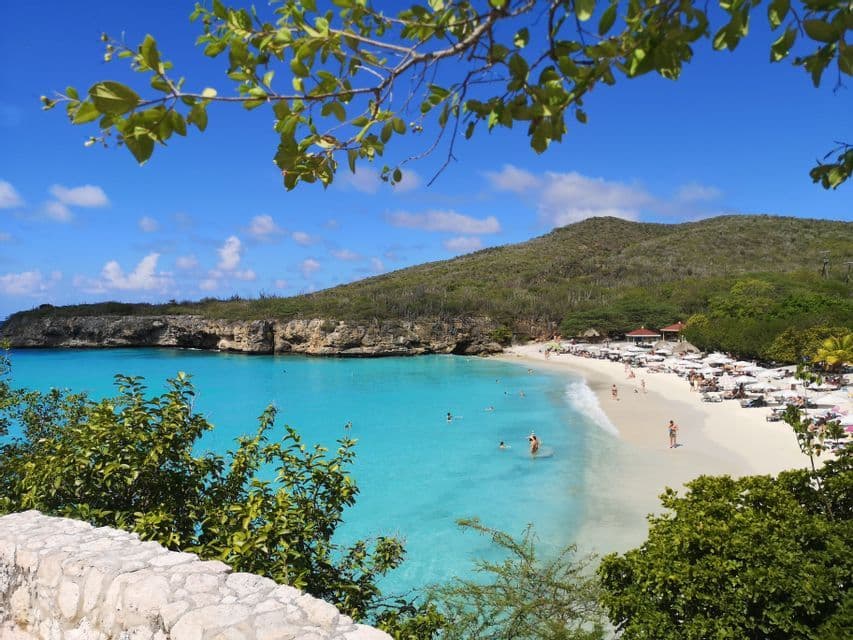 Una vista dall'alto di una caletta tropicale con persone che nuotano nell'acqua turchese e si rilassano sulla sabbia bianca sotto gli ombrelloni.