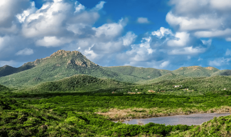 Una vista panoramica di una valle verde e lussureggiante con un fiume, che conduce a dolci colline e una montagna sotto un cielo parzialmente nuvoloso.