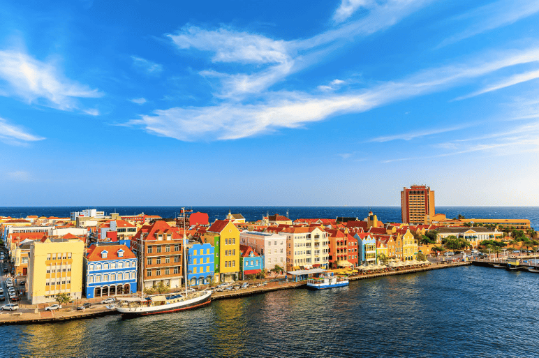 Una fila di edifici colorati si affaccia su un lungomare con barche ormeggiate, sotto un cielo azzurro brillante con nuvole sottili.