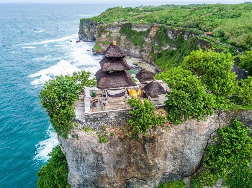 An aerial view of a traditional temple with thatched roofs perched on a sheer cliff overlooking a turquoise ocean with crashing waves.