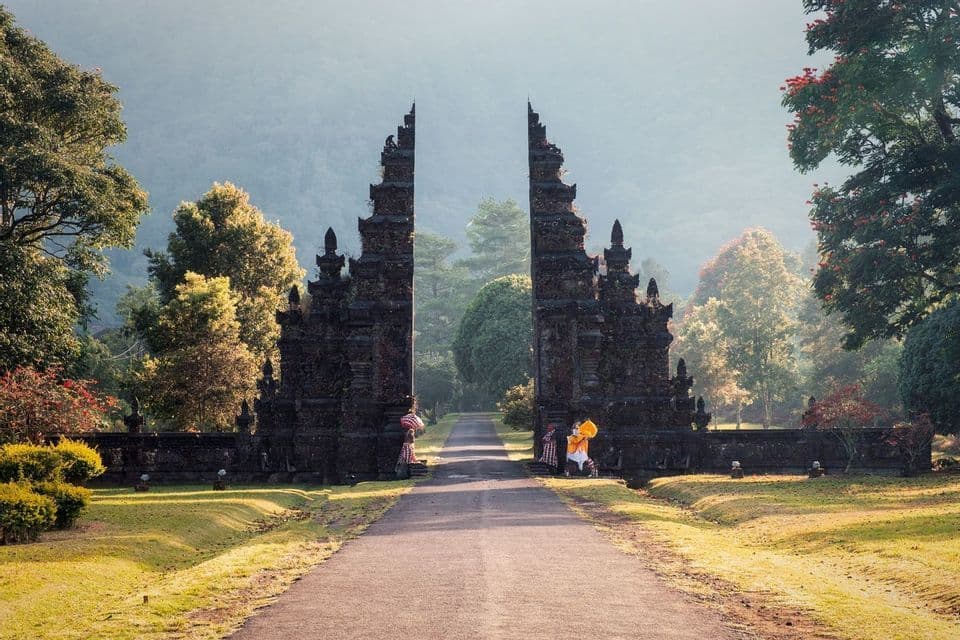 An ornate stone split-gate stands over a road in a lush landscape, with misty mountains visible in the background.