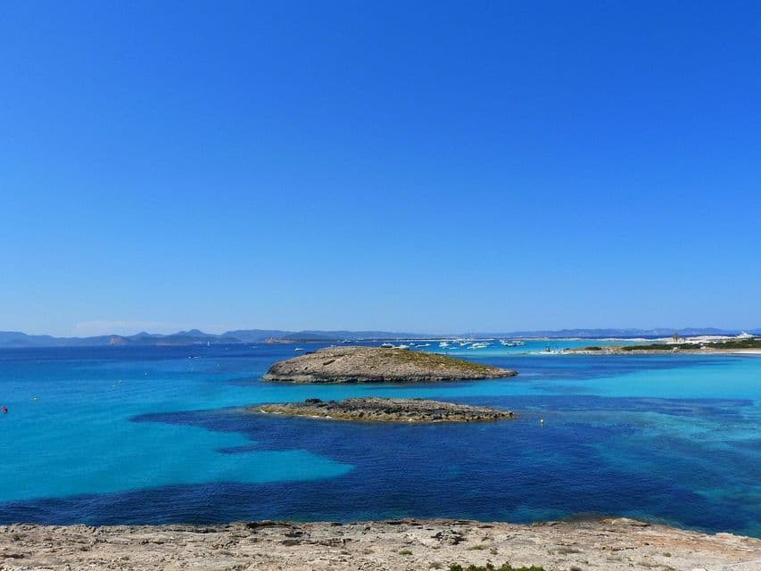 Dos islotes rocosos se asientan en aguas turquesas y azul profundo, con una costa rocosa en primer plano bajo un cielo despejado.