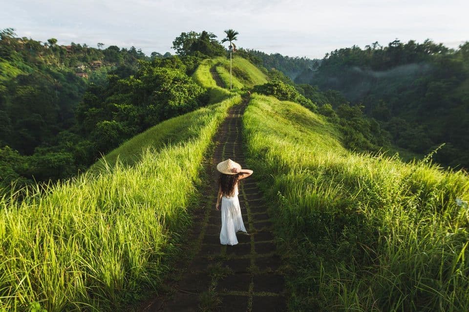 A woman wearing a conical hat and white dress walks along a stone path on a lush, green ridge.