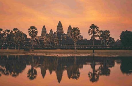 Le temple d'Angkor Wat et les palmiers environnants se reflètent dans un lac sous un ciel orange au coucher du soleil.
