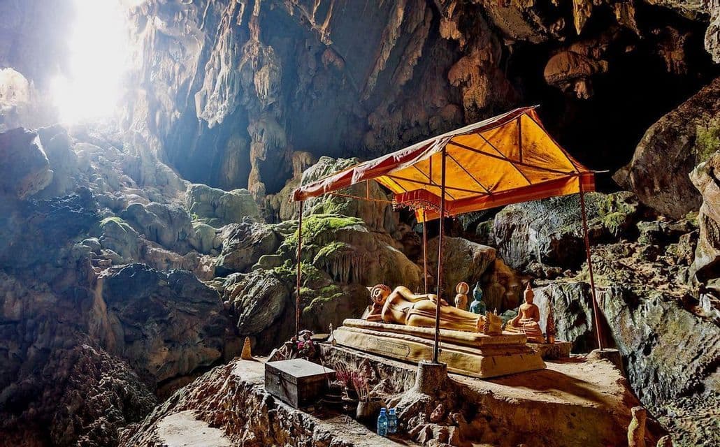 Une statue de Bouddha couché doré, sous un dais jaune, se trouve à l'intérieur d'une grande grotte rocheuse éclairée par un rayon de lumière.
