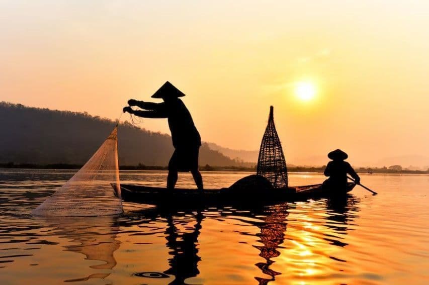 Silhouettes de deux pêcheurs aux chapeaux coniques sur une barque au lever du soleil, l'un jetant un filet de pêche dans l'eau calme.
