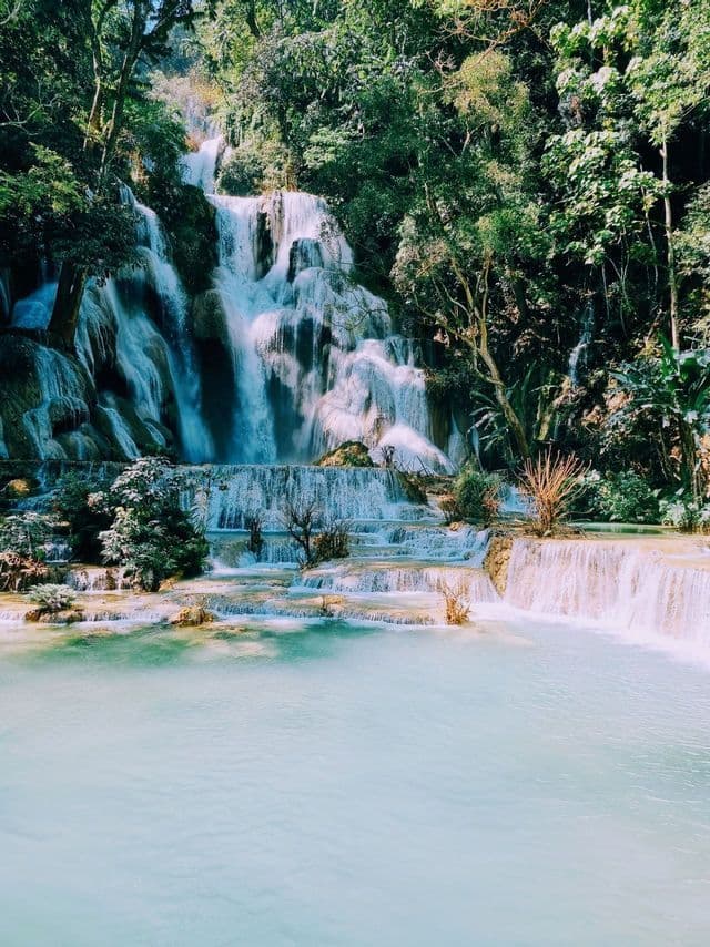 Une cascade à plusieurs niveaux dévale des formations rocheuses et se jette dans un bassin d'eau turquoise, entourée d'une forêt luxuriante et verdoyante.