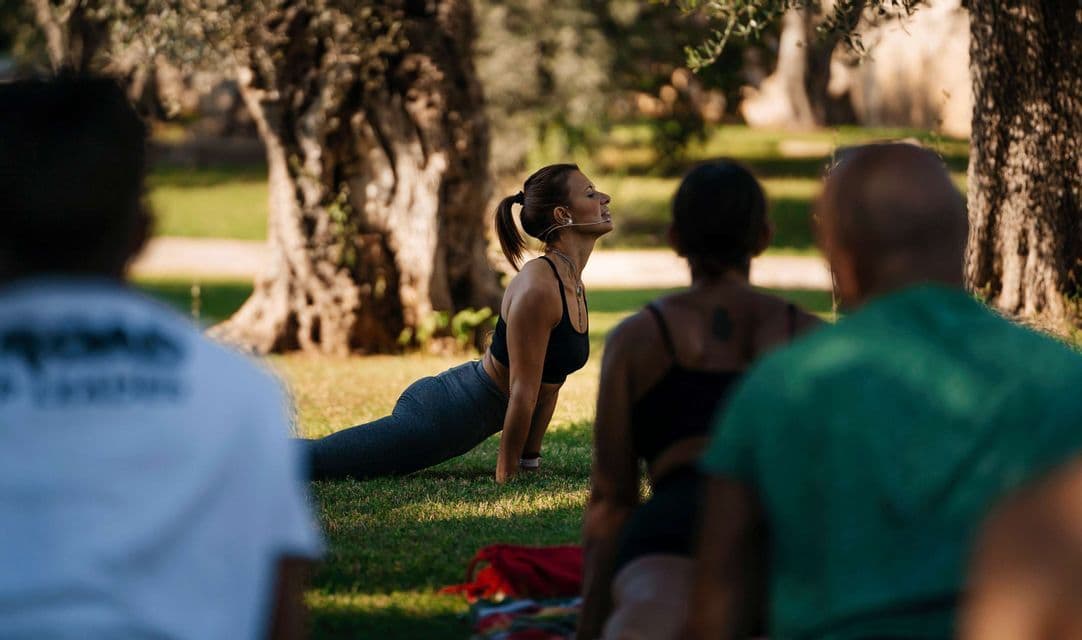 Una donna con auricolare guida un gruppo WeRoad durante una lezione di yoga all'aperto su un prato erboso, sotto gli alberi.