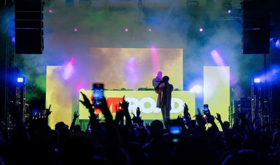 Una folla registra un concerto con i telefoni mentre un artista si esibisce su un palco con il logo 'WEROAD', un DJ e luci colorate.