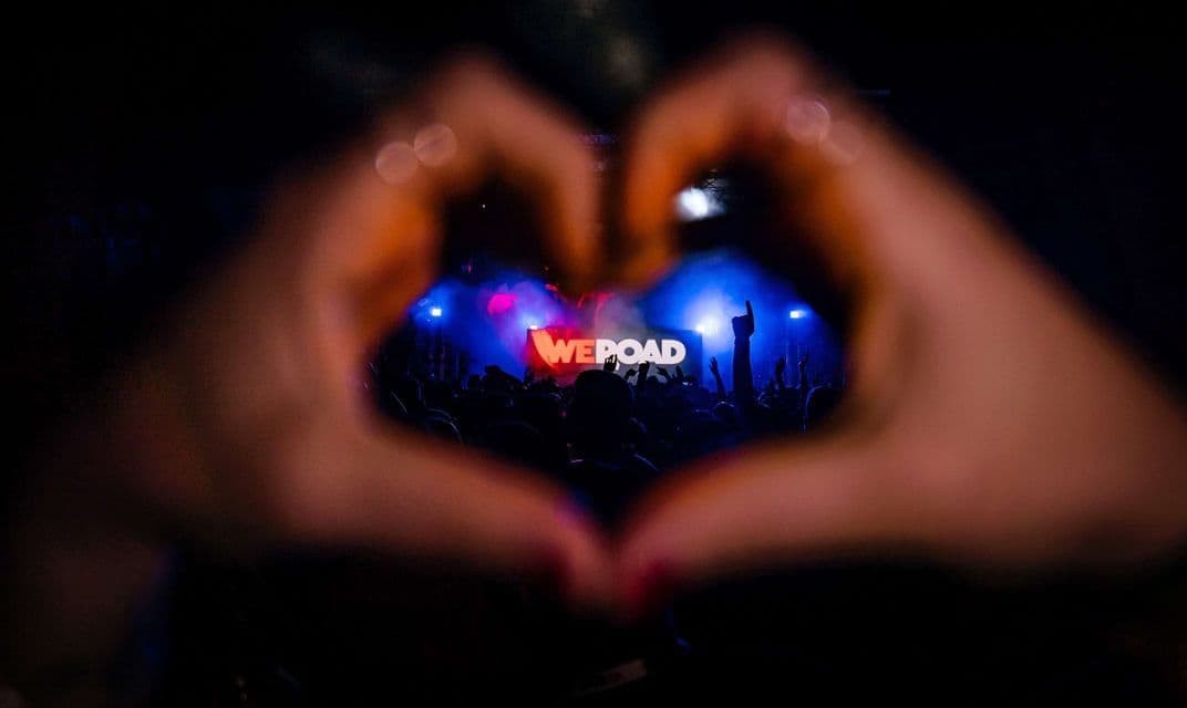 Mani a forma di cuore incorniciano la vista di un palco di concerto affollato con il logo WeRoad illuminato di blu e rosso.