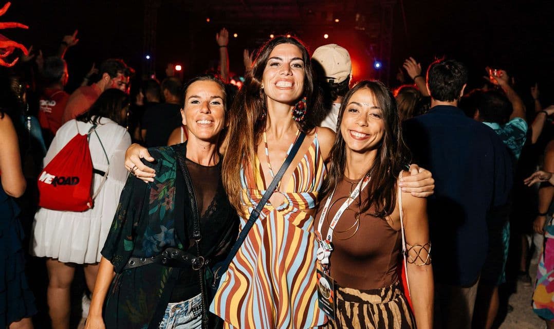 Tre donne in un viaggio di gruppo WeRoad sorridono e posano per una foto in mezzo alla folla a una festa notturna.