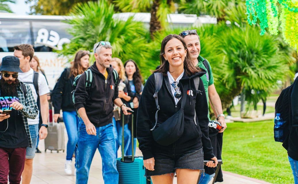 Un gruppo WeRoad in viaggio cammina all'aperto con i bagagli, mentre una donna al centro sorride alla fotocamera con un autobus e palme sullo sfondo.