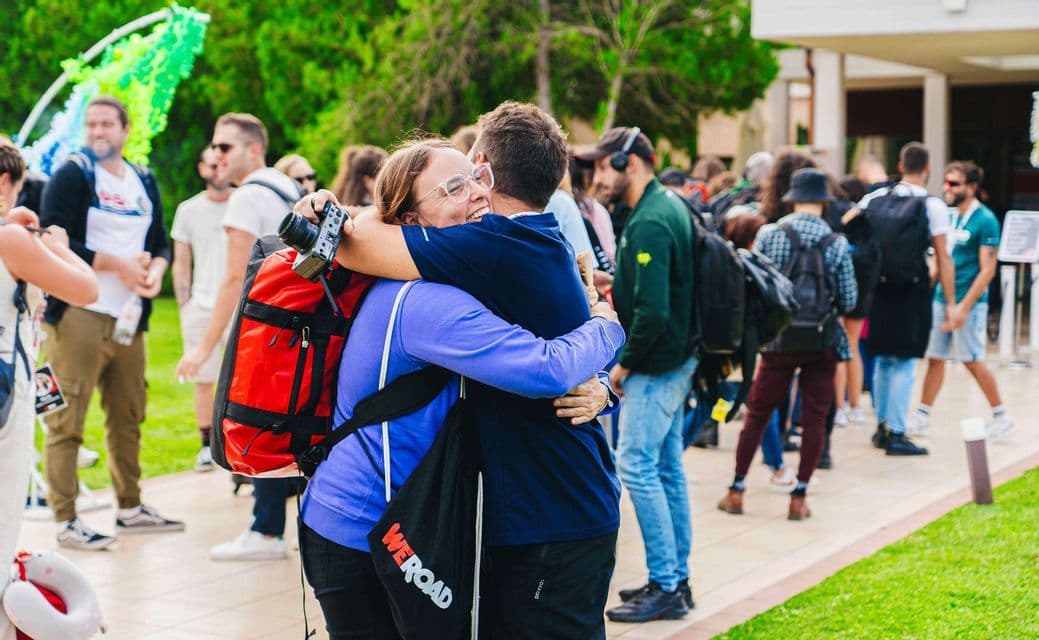 Una donna sorridente con una macchina fotografica abbraccia un'altra persona durante un raduno di gruppo WeRoad all'aperto.