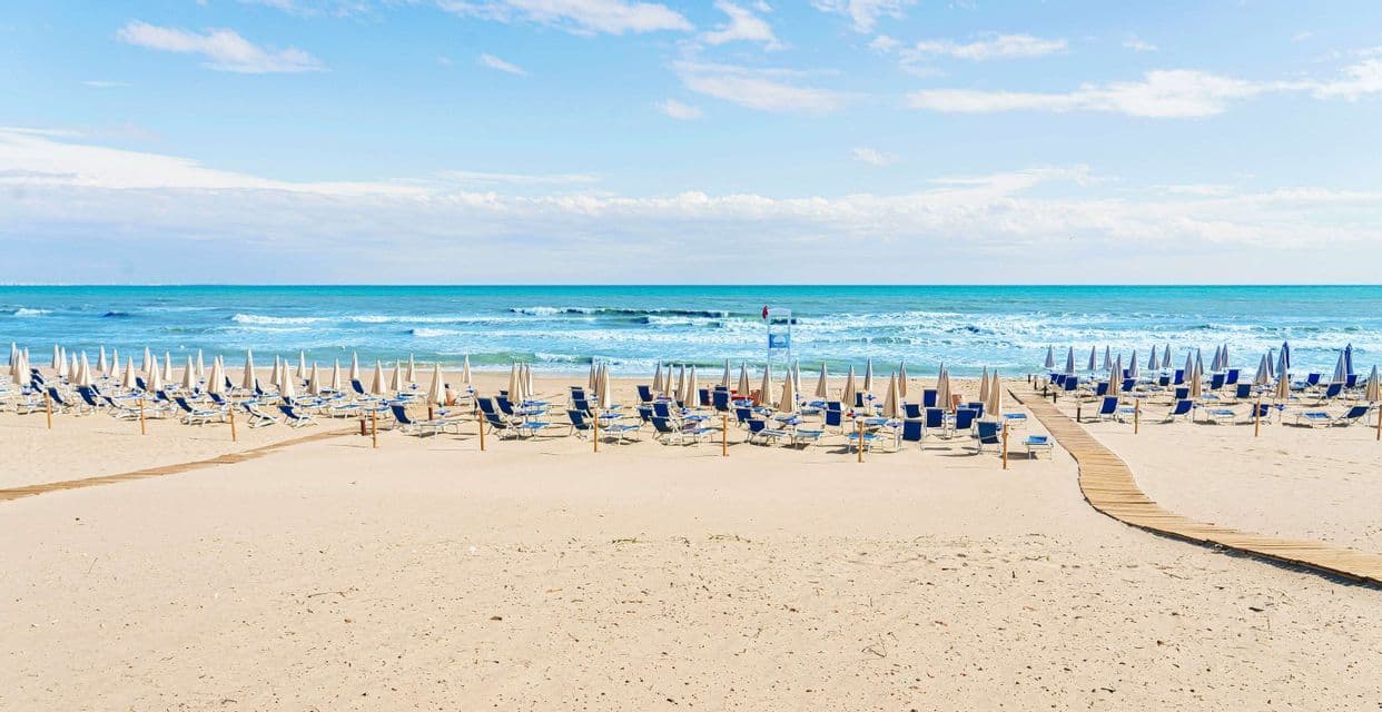 File di sdraio vuote e ombrelloni chiusi sono disposte su una spiaggia sabbiosa con passerelle in legno che conducono verso l'oceano turchese.