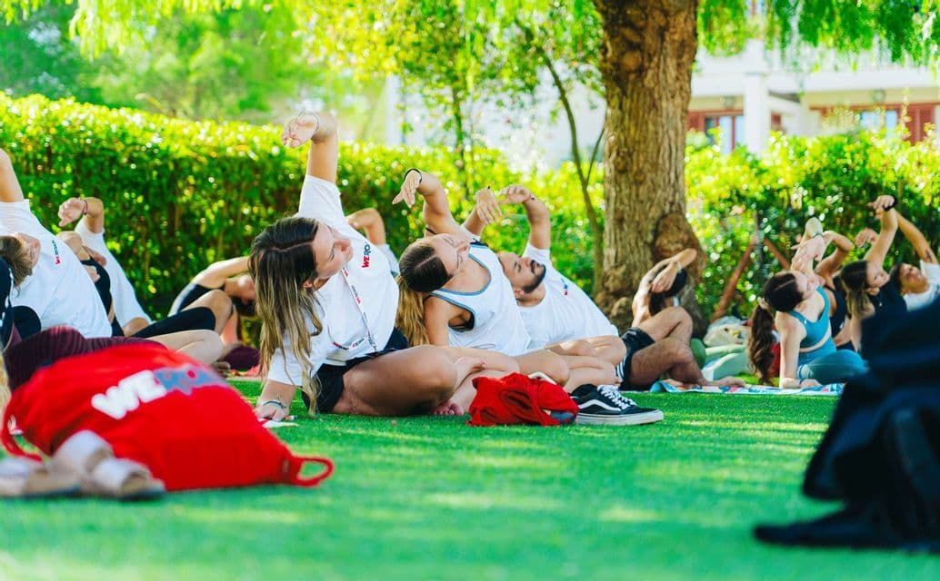Un viaggio di gruppo WeRoad partecipa a una sessione di yoga all'aperto, facendo stretching insieme su un prato verde e soleggiato sotto gli alberi.