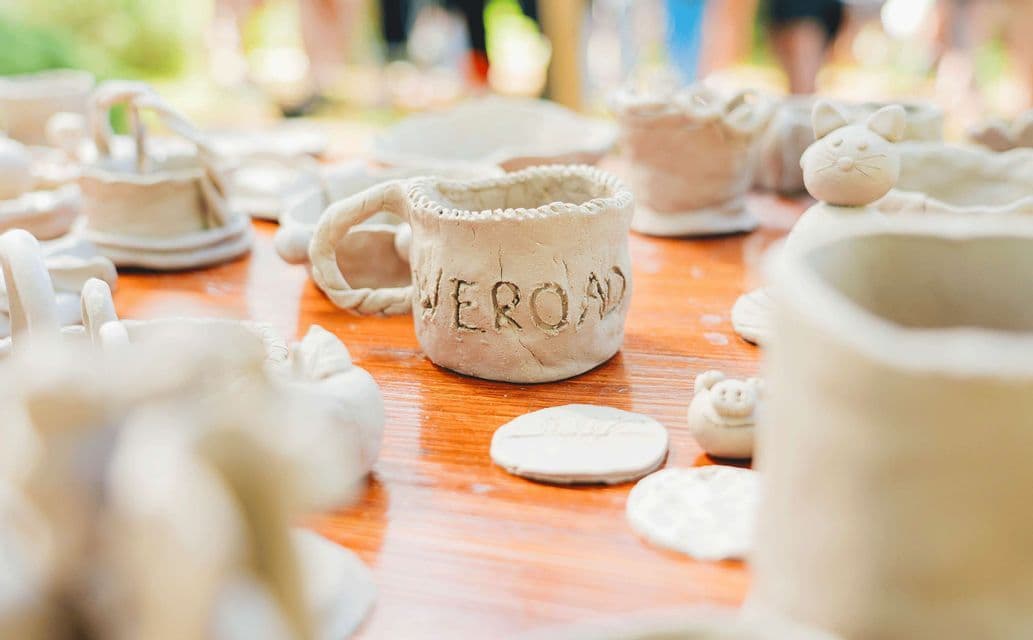 Una collezione di creazioni artigianali in argilla cruda su un tavolo di legno, inclusa una tazza con la parola 'WEROAD' intagliata sul lato.