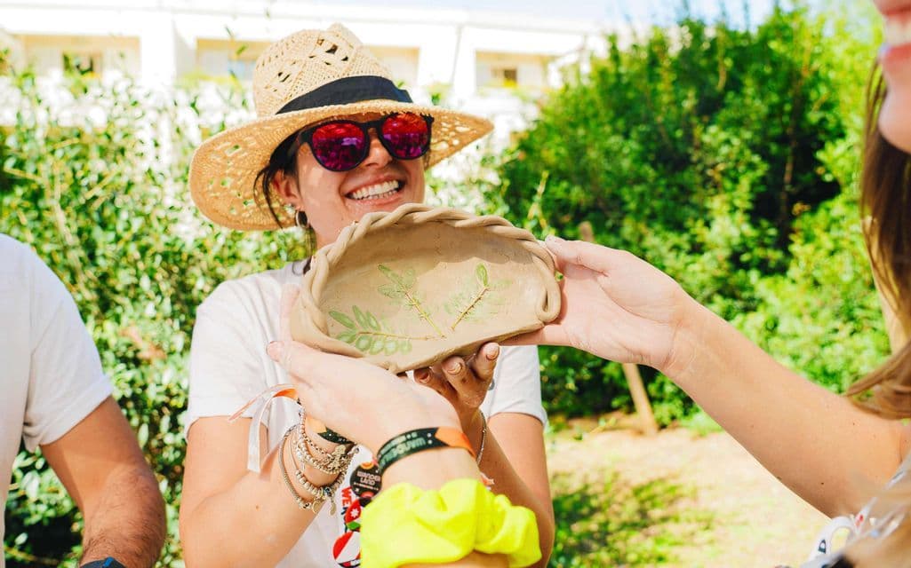 Una donna sorridente con un cappello di paglia mostra un piatto di argilla fatto a mano con impronte di foglie durante un'attività di viaggio di gruppo WeRoad.