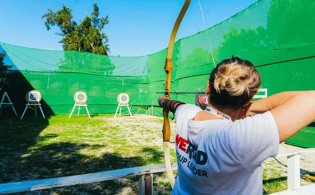 Una capogruppo WeRoad con i capelli biondi raccolti in uno chignon mira con arco e freccia a dei bersagli su un campo di tiro con l'arco all'aperto.