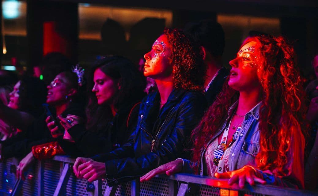 Un viaggio di gruppo WeRoad di donne con brillantini sui volti che guardano un concerto da dietro una transenna sotto luci rosse e blu.