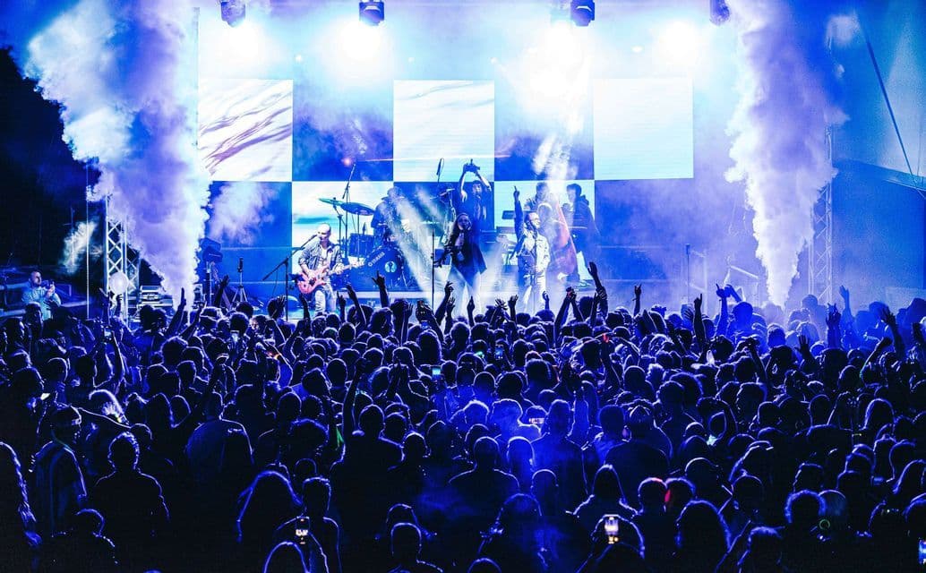 Una band si esibisce su un palco con luci blu intense e fumo davanti a una grande folla con le braccia alzate.