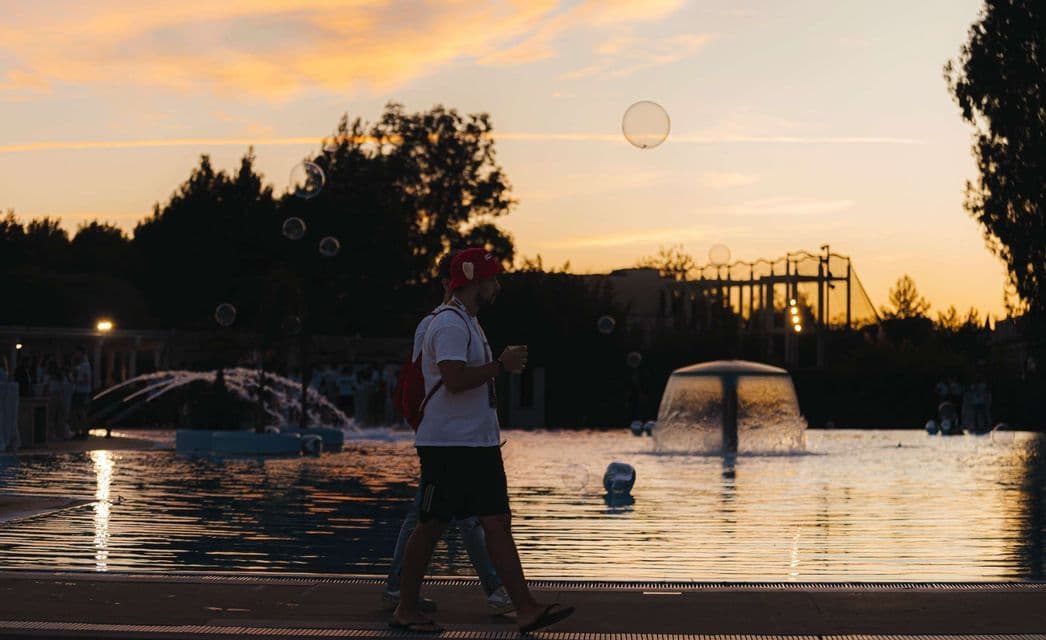 Un uomo con un berretto rosso cammina a bordo piscina con fontane al tramonto, mentre bolle di sapone gli fluttuano intorno.