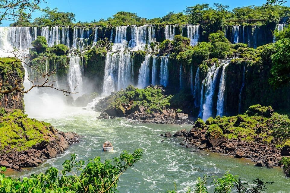 Un bateau de tourisme navigue sur une rivière au pied d'une immense cascade étagée, entourée de falaises verdoyantes et luxuriantes.