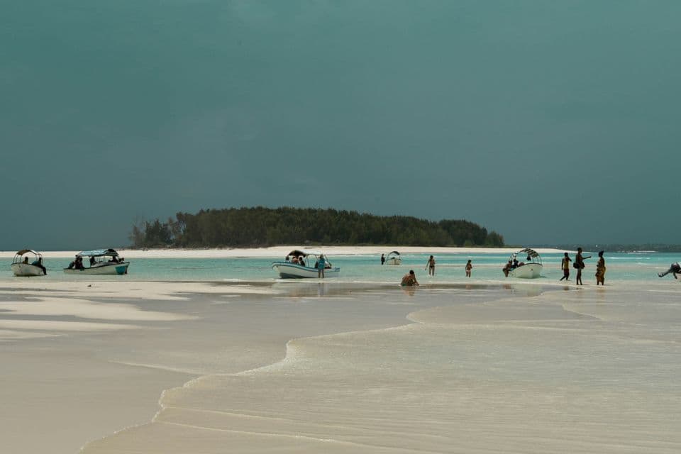 Un viaggio di gruppo WeRoad che guada in acque basse e turchesi con piccole barche su una spiaggia di sabbia bianca sotto un cielo coperto.