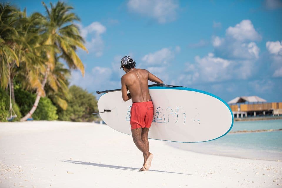 Una persona di spalle, in pantaloncini rossi e berretto, porta un paddleboard mentre cammina su una spiaggia di sabbia bianca con palme.