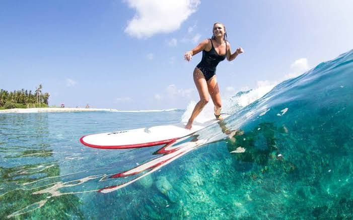 Una donna in costume nero fa surf su un'onda in acqua cristallina turchese, con una spiaggia tropicale e palme sullo sfondo.
