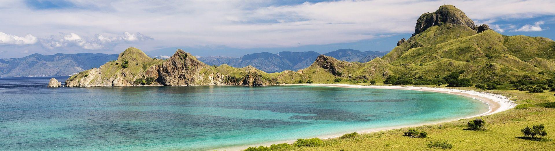 Une vue panoramique sur une baie aux eaux turquoise, une plage de sable blanc et des collines verdoyantes escarpées sous un ciel partiellement nuageux.