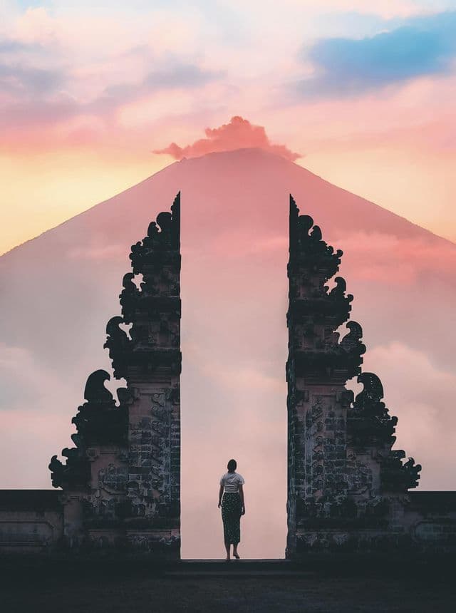 Une personne vue de dos se tient entre deux piliers de pierre ouvragés, encadrant la vue d'un volcan lointain au coucher du soleil.