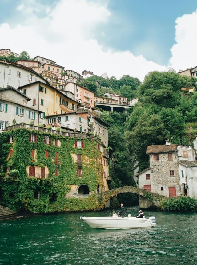 Un motoscafo bianco con due persone naviga accanto a un ripido villaggio collinare con un edificio coperto d'edera e un piccolo ponte di pietra.