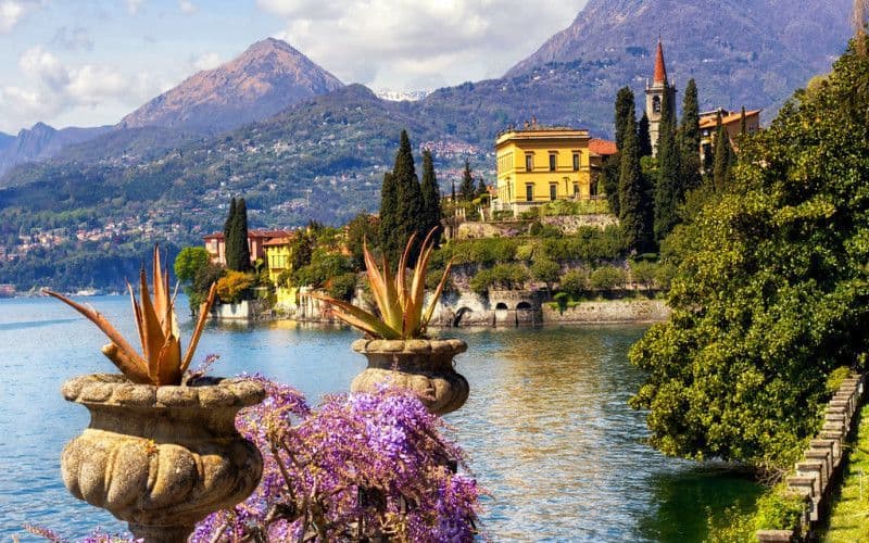 Una villa gialla e un campanile sulla sponda di un lago, ai piedi delle montagne, con fiori viola e vasi di pietra in primo piano.