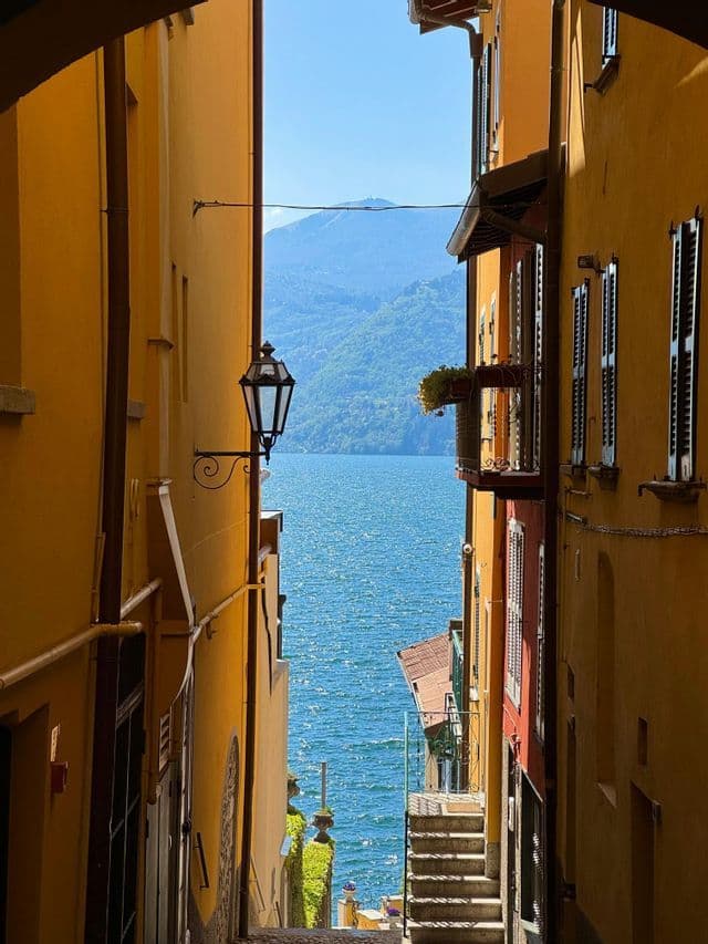 Uno stretto vicolo tra edifici gialli incornicia la vista di un lago azzurro brillante e di lontane montagne verdi sotto un cielo sereno.