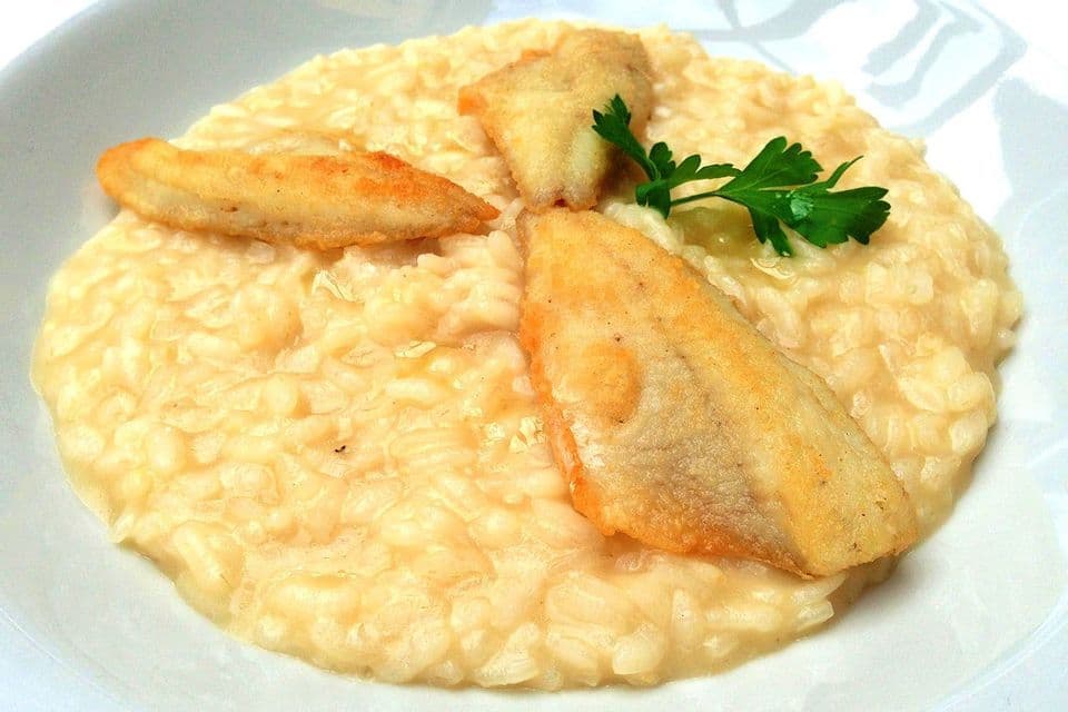 Un primo piano di risotto cremoso guarnito con tre filetti di pesce dorati e fritti e un rametto di prezzemolo in una ciotola bianca.